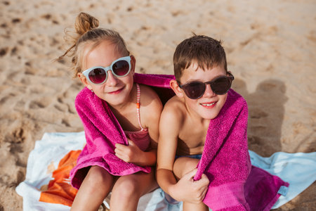 Siblings sitting on beach, wrapped in towel after swim in sea. Concept of beach summer vacation with kids.の写真素材