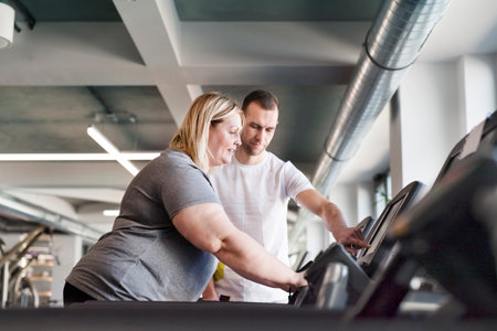 Overweight woman exercising on treadmill in gym. Personal trainer helping her.