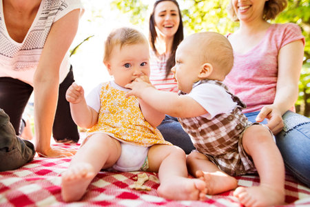 Mothers and babies enjoying group picnic outdoor in park, sitting on picnic blanket and preparing food and drinks.の写真素材