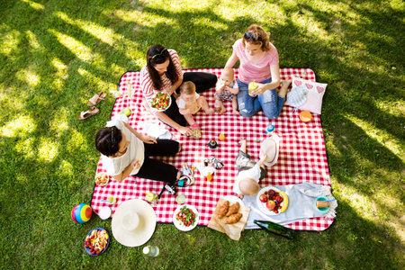 Mothers and babies enjoying group picnic outdoor in park, sitting on picnic blanket and preparing food and drinks.の写真素材