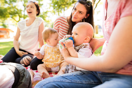 Mothers and babies enjoying group picnic outdoor in park, sitting on picnic blanket and preparing food and drinks.の写真素材