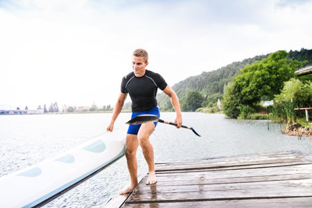 Young canoeist carry canoe and paddle, going into water, walking on wooden dock. Concept of canoeing as dynamic and adventurous sportの写真素材