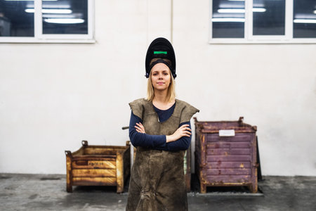 Beautiful blonde woman works as a welder in workshop, operating welding machine, wearing protective clothing and a welding mask.の写真素材