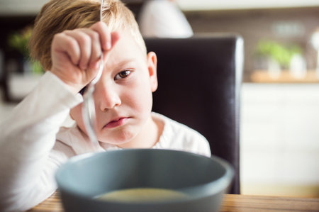 Little boy doesnt like lunch, doesnt want to eat. Sitting at table with serious expression, holding spoon, refusing to eat.の写真素材