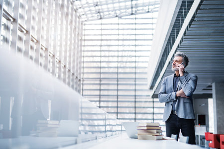 Handsome businessman working on laptop, searching for informations in book and making phone call. Traditional information source in modern office space.の写真素材