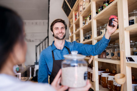 Handsome shop assistant serving customer in package-free store using reusable containers. Zero waste...