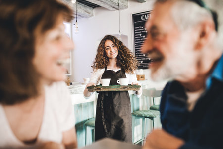 Beautiful female barista working in coffee shop, serving customers, elderly couple. University...