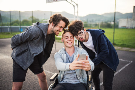 Young man in wheelchair spending free time outdoor with friends, taking selfie with smartphone. Male friendship.の写真素材