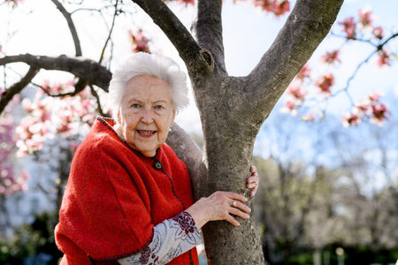 Portrait of elderly woman standing by magnolia tree, in park, having relaxing moment. Grandmother...
