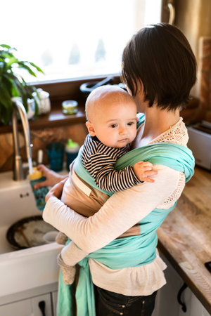 Mother holding small baby while cleaning dishes, carrying him around house in baby wrap. Household...