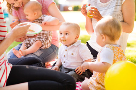 Mothers and babies enjoying group picnic outdoor in park, sitting on picnic blanket and preparing food and drinks.の写真素材