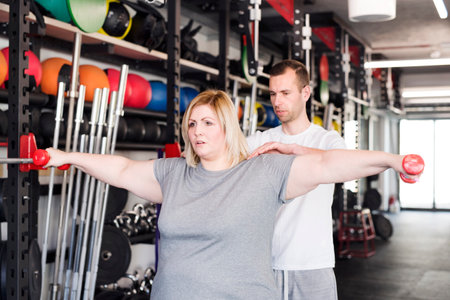 Overweight woman exercising in gym, using dumbbells. Personal trainer couching her and helping her.の写真素材