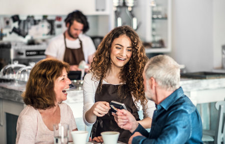 Beautiful female barista working in coffee shop, serving customers, elderly couple. University...