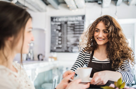 Beautiful female barista working in coffee shop, serving coffee to female customer. University...