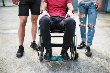 Close up of legs of young man sitting in wheelchair, friends standing by him. Male friendship.の写真素材