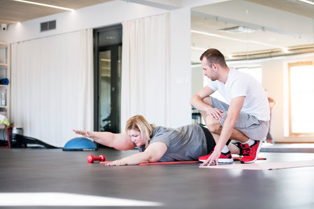 Overweight woman exercising on gym mat in gym. Personal trainer couching her and helping her.