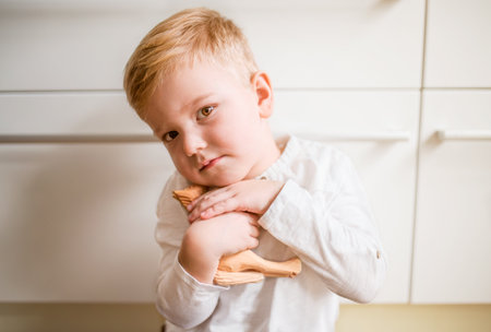 Portrait of a cute boy sitting on the floor in the kitchen, holding wooden horse toy.