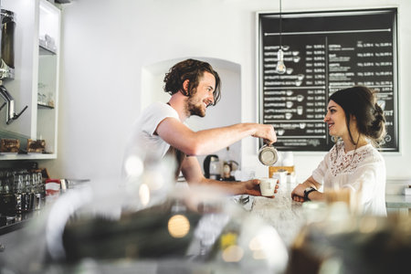 Handsome barista working in coffee shop, serving female customer, preparing coffee drink. University...