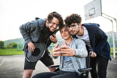 Young man in wheelchair spending free time outdoor with friends, taking selfie, having fun in city. Male friendship.の写真素材