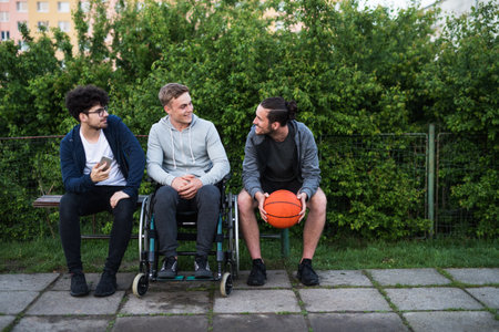 Young man in wheelchair spending free time outdoor with friends, having fun in city. Male...