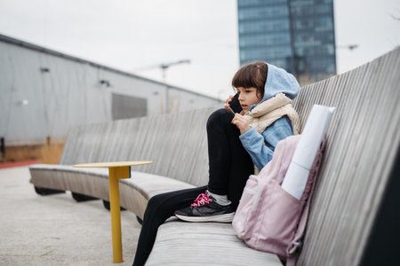 Sad girl phone calling, sitting in front of school building, waiting for parents. Young schoolgirl...