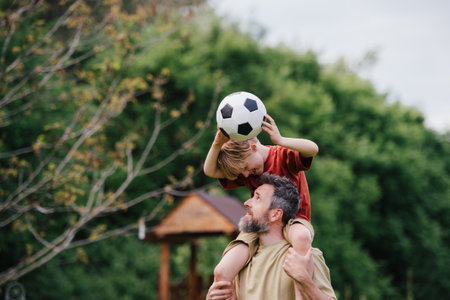 Dad having fun with young son, playing football, carrying boy on shoulders. Playing on a lawn in front of their house. Fatherhood and Fathers Day.の写真素材