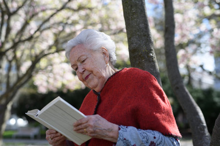 Beautiful elderly woman reading book outdoor, in park, having relaxing moment. Grandmother reading...
