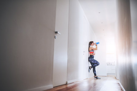 Beautiful redhead woman drinking water, resting after home fitness routine. Home workout on gym mt in living room, sporty woman wearing sportswear.の写真素材