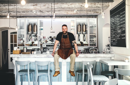 Handsome male barista working in coffee shop, sitting on counter. Bartender in apron preparing...