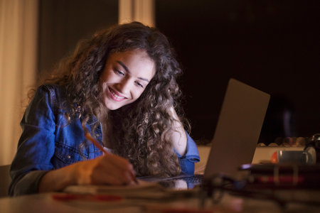 A beautiful university student having evening online class, e-learning sitting at home. Online...