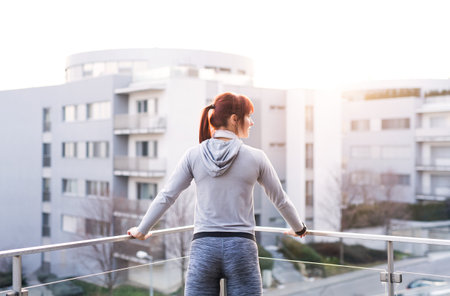 Rear view of beautiful redhead woman after fitness routine, standing on balcony, resting. Home workout, sporty woman wearing sportswear.の写真素材