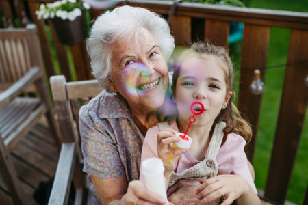 Granddaughter blowing bubbles with elderly grandma. Senior lady spending time with young girl, enjoying together time.の写真素材