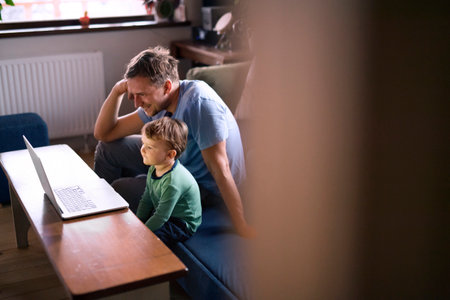 Dad and boy watching cartoons on laptop, sitting on sofa in living room, having great time together.の写真素材