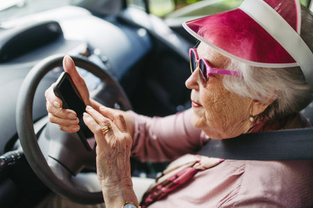 Happy senior woman driving car alone, holding smartphone and scrolling. Safe driving for elderly...