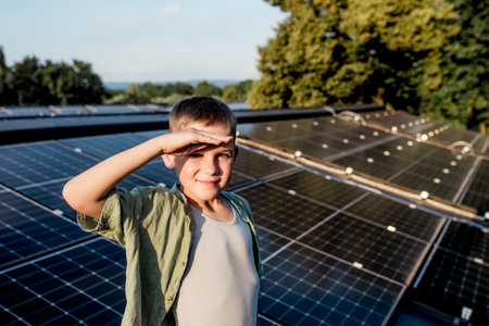 Cute boy on roof with solar panels, hand shielding eyes from summer sun. Rooftop solar or photovoltaic system. Sustainable future for next generation.の写真素材