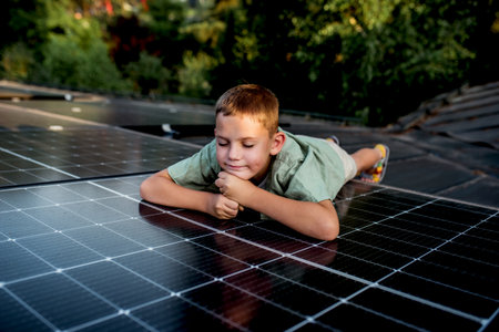 Boy lying on belly on roof with solar panels, looking at surrounding nature. Rooftop solar or...