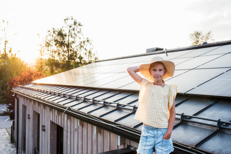 Girl standing in front of roof with solar panels during sunset, smiling and holding hat. Rooftop solar or photovoltaic system. Sustainable future for next generation.の写真素材