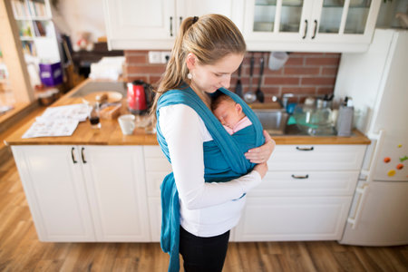 Mother holding small baby, carry her in baby sling, wrap around the house. Unconditional paternal...