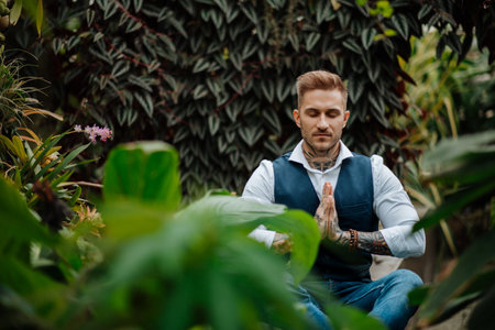 Handsome man with tattoo meditating in botanical garden, in the middle of lush green foliage. Taking...