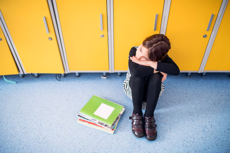Portrait of schoolgirl sitting by lockers. Student girl with bag in hallway, after day in elementary schoolの写真素材