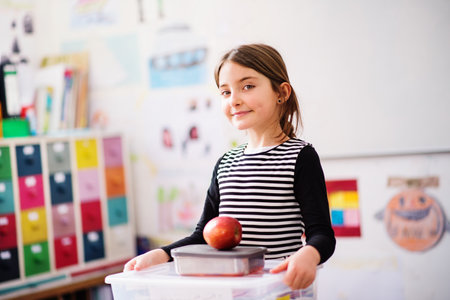 Portrait of schoolgirl, pupil sitting at desk in classroom at the elementary school. Student girl holding apple during snack time, breakの写真素材