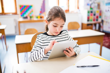 Portrait of cute schoolgirl sitting at desk using tablet during class, learning with technology. Student girl in classroom at the elementary schoolの写真素材