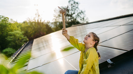 Cute girl on roof with solar panels, holding model of wind turbine. Rooftop solar or photovoltaic system. Sustainable future for next generation.の写真素材