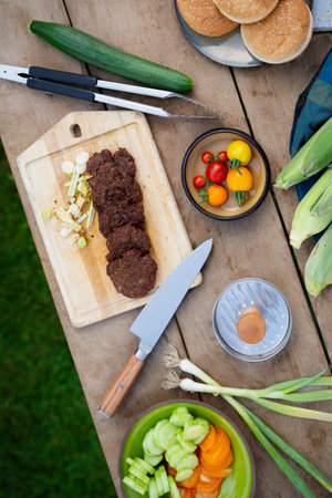 Top view of cutting board with sharp knife, chopped vegetables and grilled burger patties. Preparing food for an outdoor barbecue. grilling setup.の写真素材