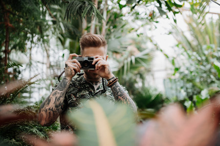 Handsome man with tattoos standing in botanical garden, in the middle of lush green foliage, holding camera and taking photography.の写真素材
