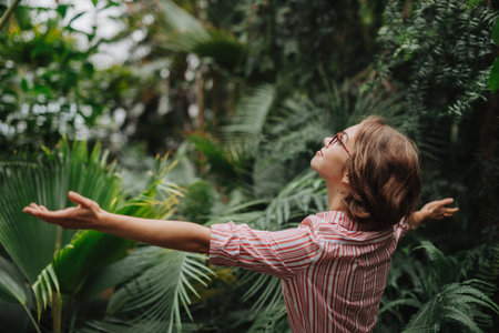 Beautiful woman standing in botanical garden, in the middle of lush green foliage, arms open. Rear view.の写真素材