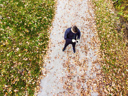 Aerial view of a runner standing autumn park on path with fallen leaves, holding smartphone, checking training performance on smartwatch. Morning running training.の写真素材
