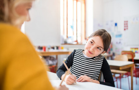 Hardworking teacher learning with young schoolgirl in classroom, sitting by desk. One to one lesson.