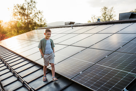 Boy walking on roof with solar panels during sunset, smiling, hands in pockets. Rooftop solar or...