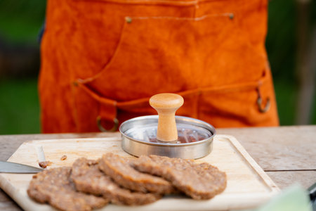 Man in apron preparing meat for hamburgers, standing outdoors. Using burger patty press to shape...